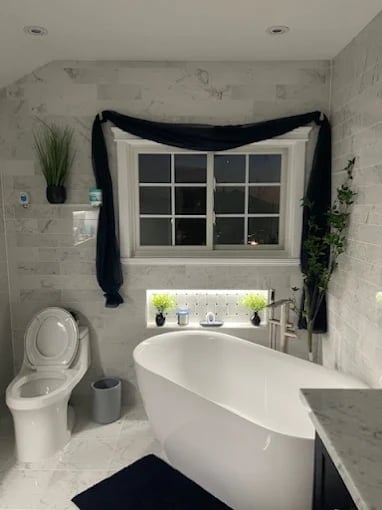 Modern bathroom with white free-standing tub, gridded window with navy curtains, toilet, and potted plants on windowsill